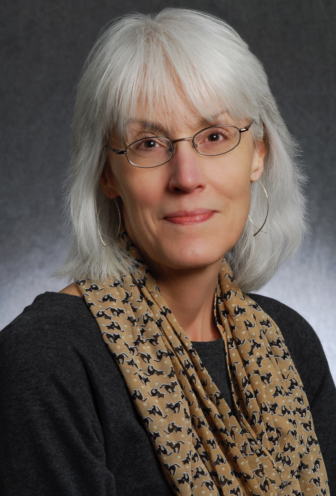 Portrait of a woman with shoulder-length gray hair wearing glasses, hoop earrings, and a patterned scarf against a neutral background.
