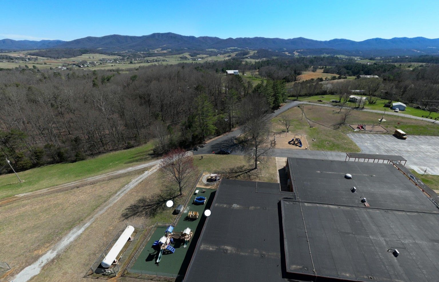 Aerial view of a rural school campus with playground equipment, parking areas, and surrounding fields, set against rolling farmland and distant mountain ridges under a clear blue sky.