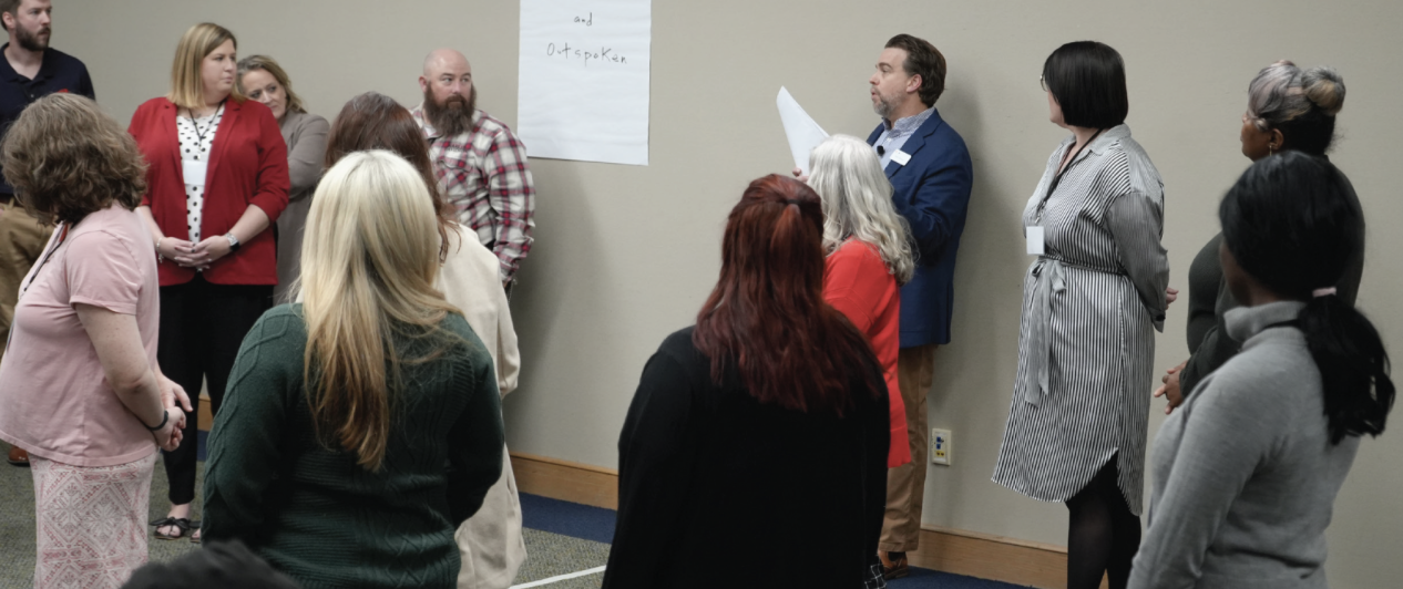 Several people standing in a training room, listening to a person speaking.