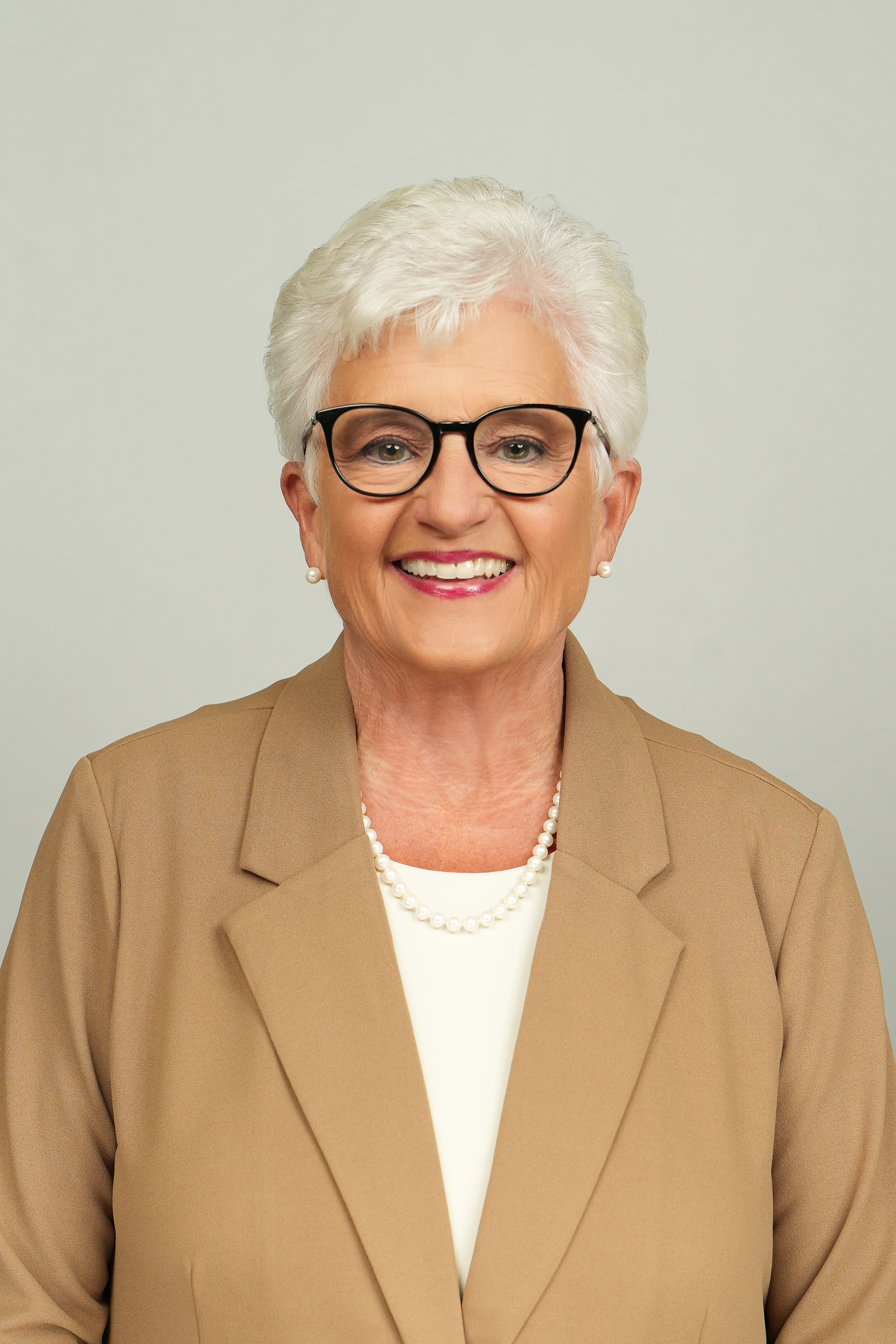Professional headshot of a smiling person with short white hair and black glasses, wearing a tan blazer.