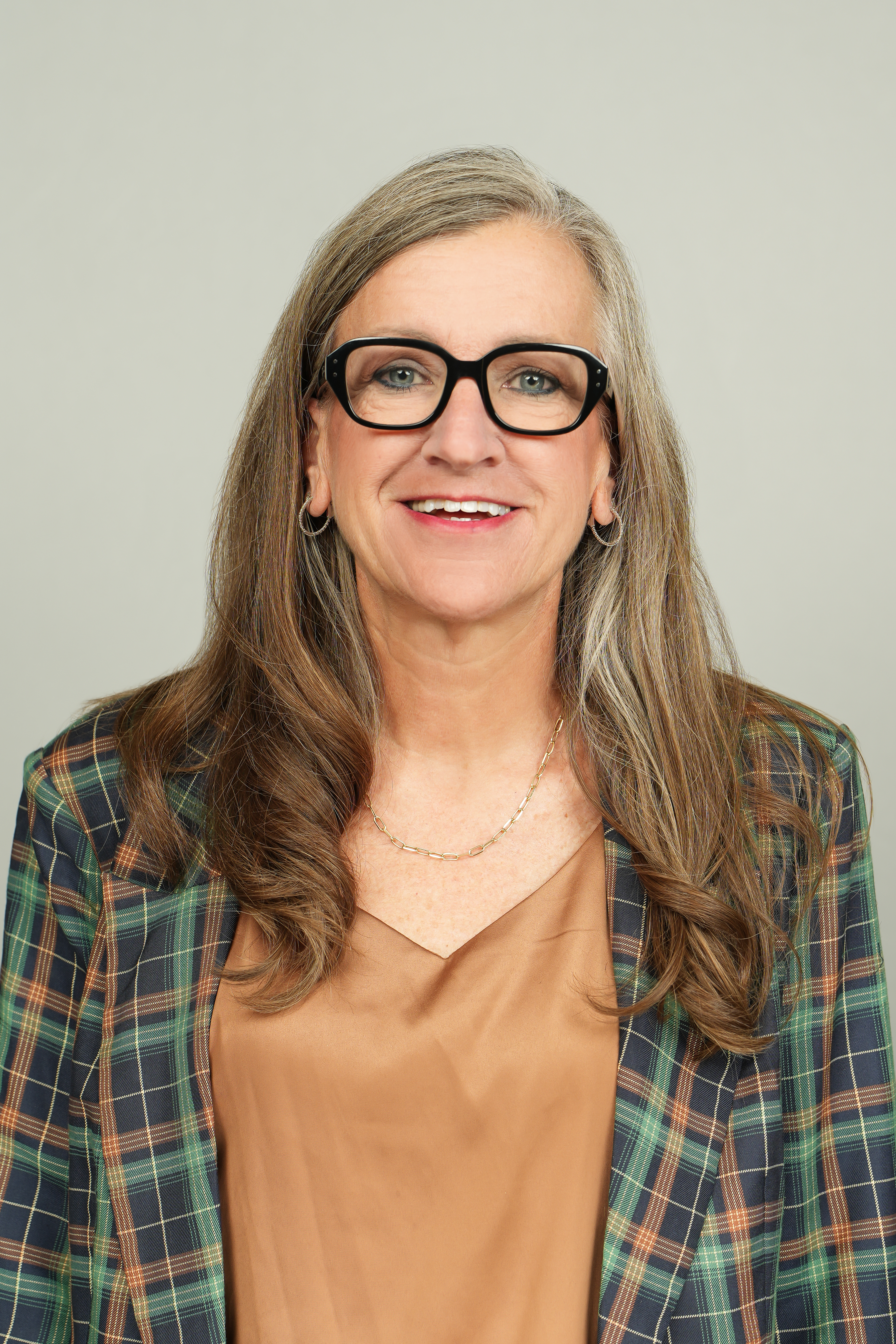Professional headshot of a smiling person with long brown-and-gray hair and black glasses, wearing a plaid blazer.