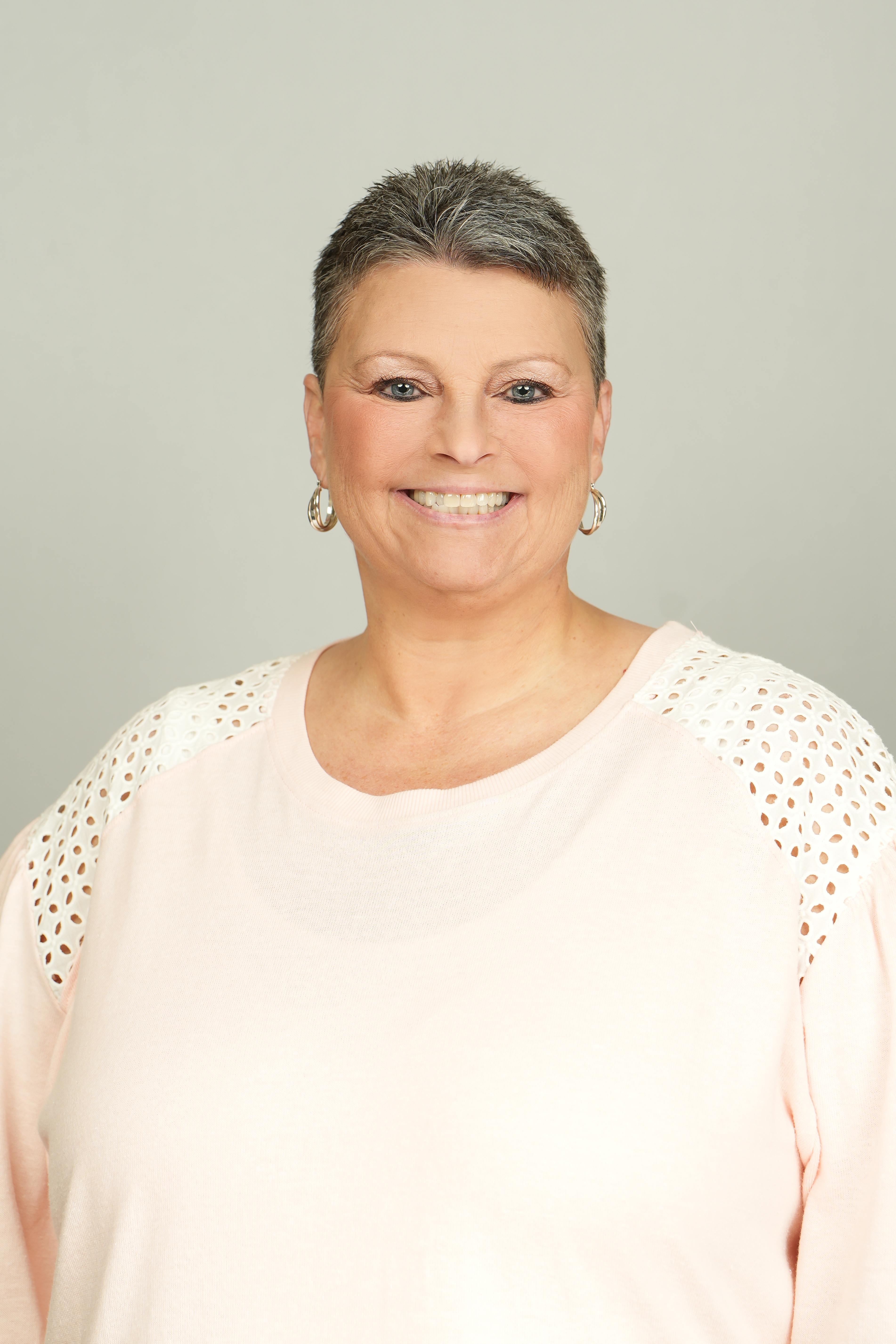 Professional headshot of a smiling person with short salt-and-pepper hair, wearing a pale pink top.
