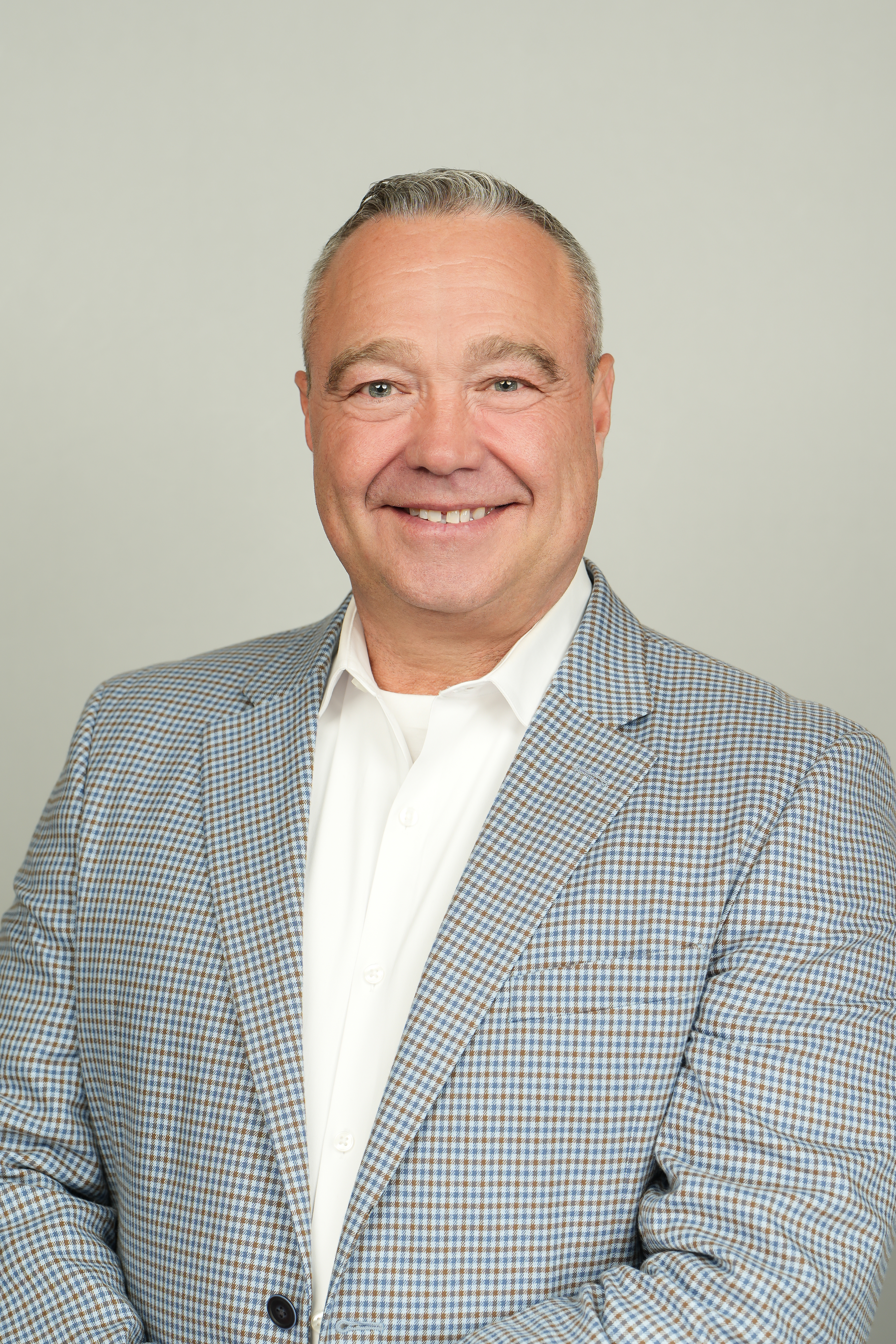 Professional headshot of a smiling person with short gray hair, wearing a blue-and-brown checked blazer.