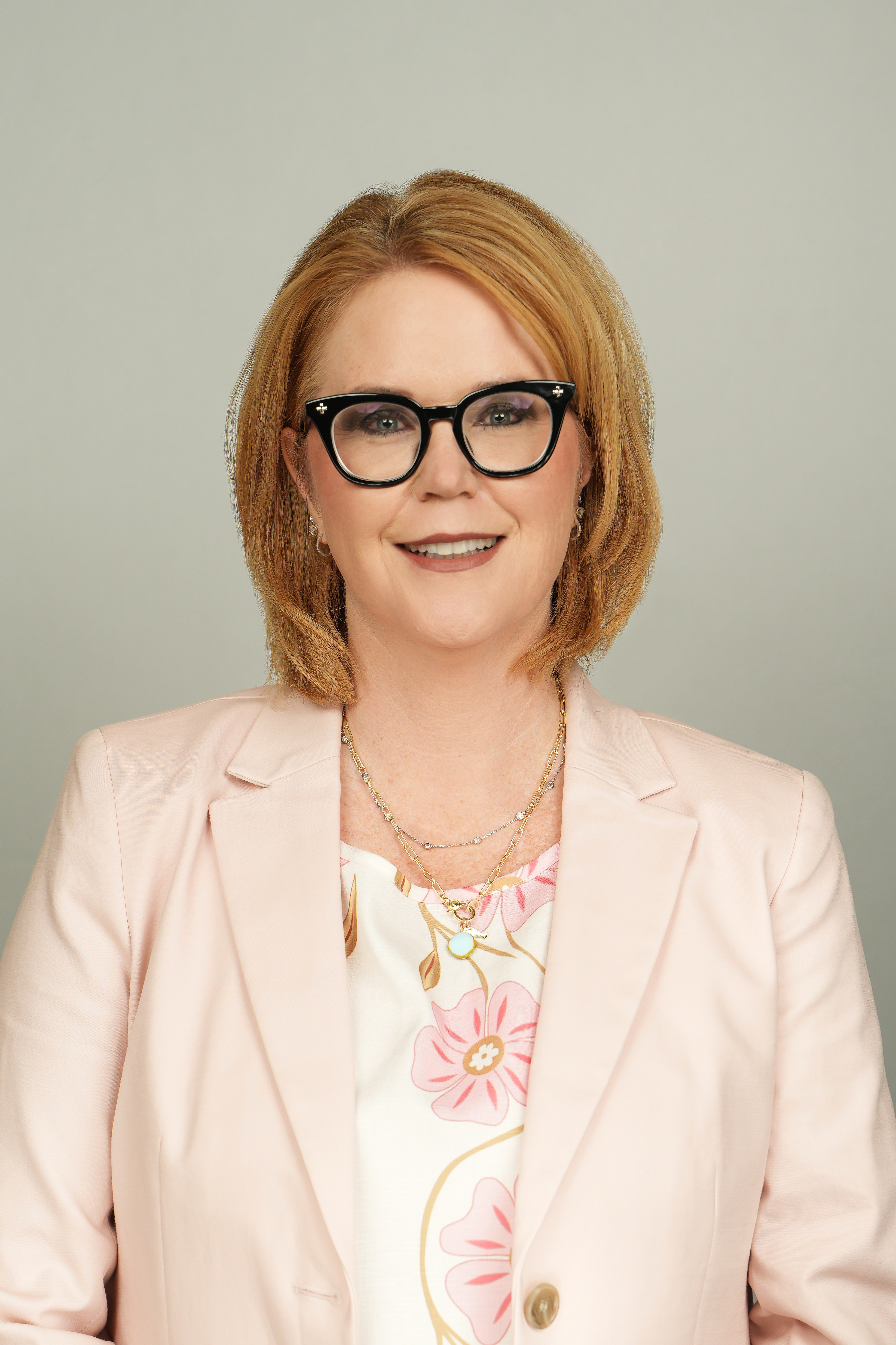 Professional headshot of a smiling person with short auburn hair and black glasses, wearing a pale pink blazer.