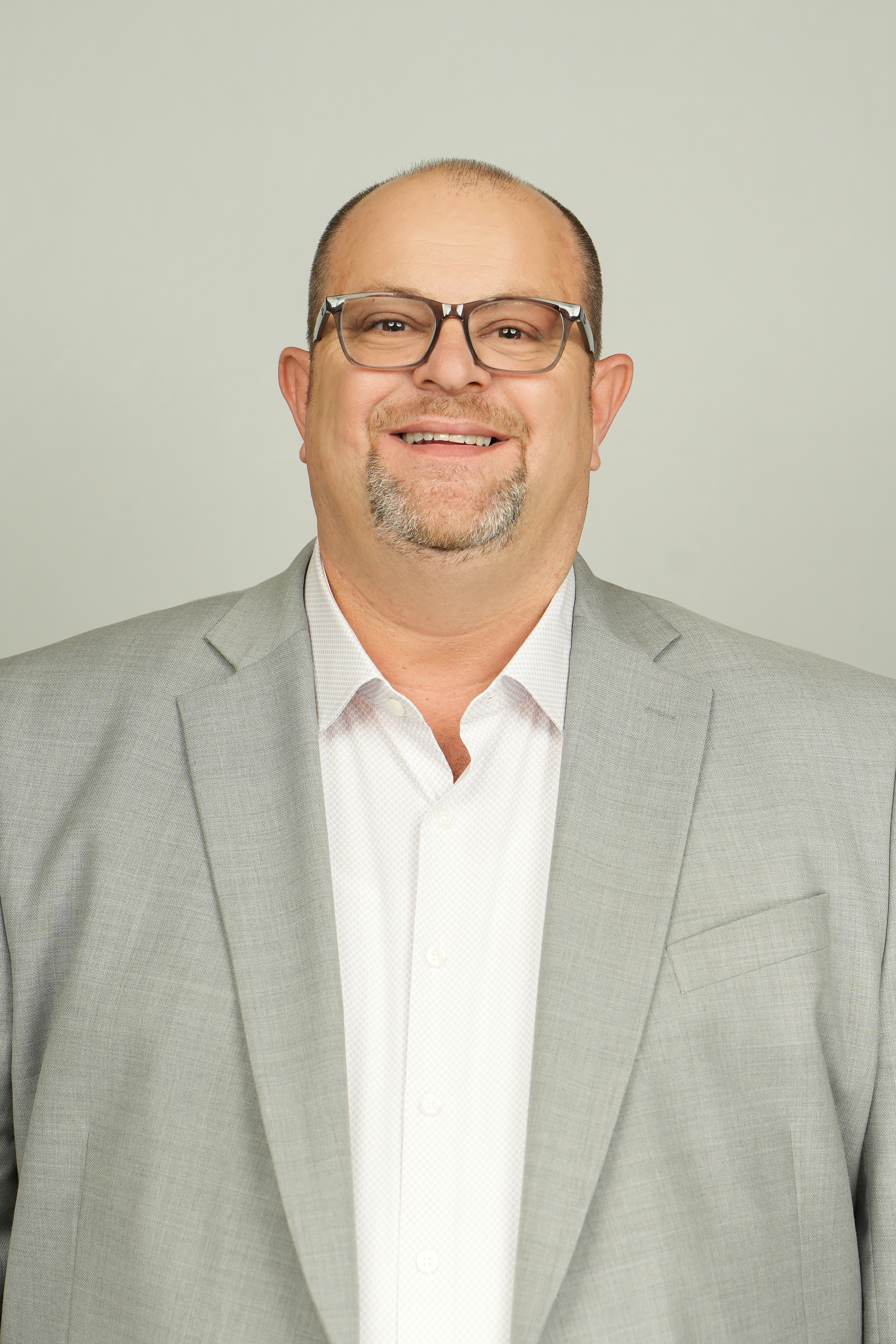 Professional headshot of a smiling person with glasses and a goatee, wearing a light gray suit jacket.