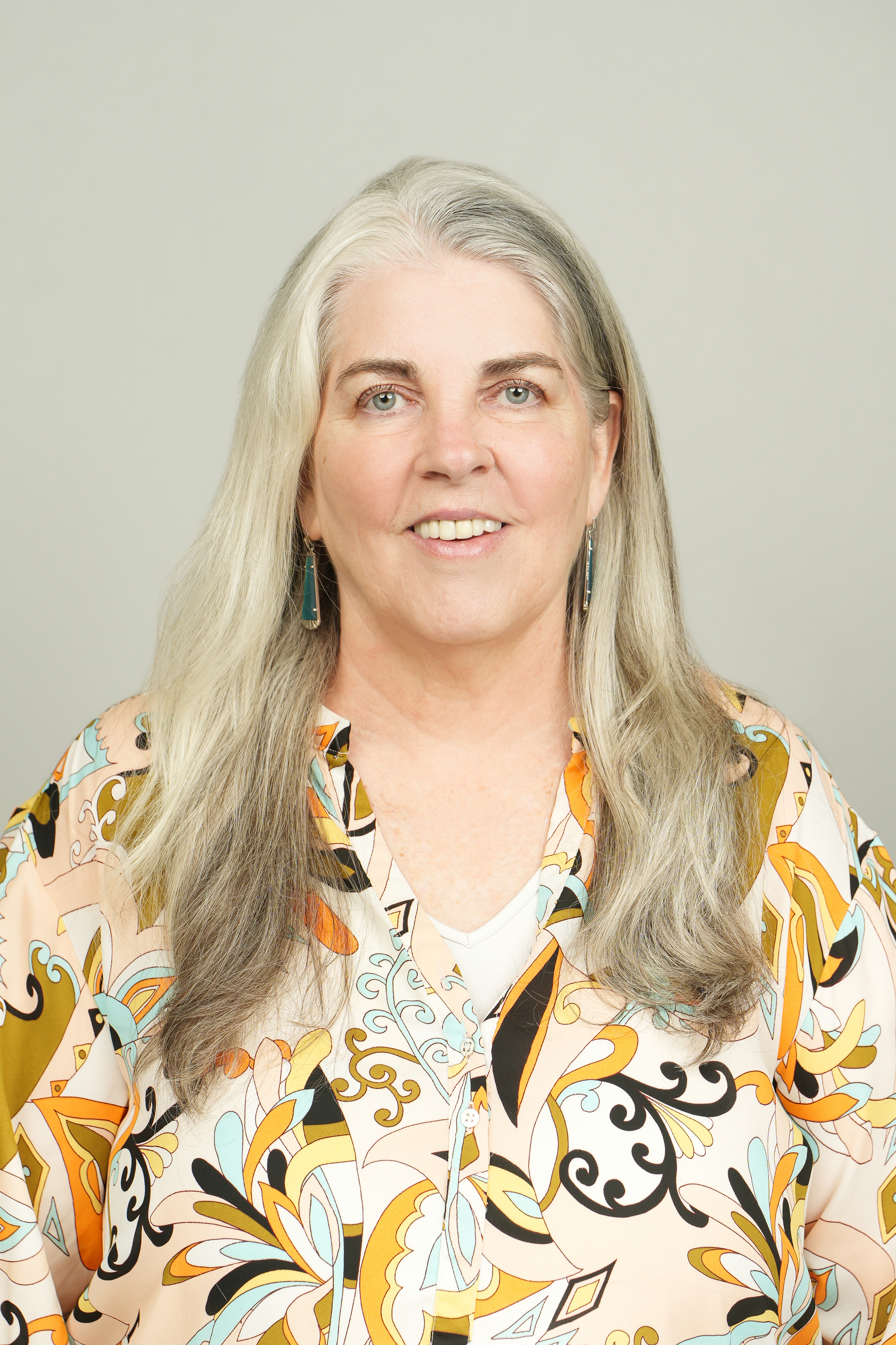 Professional headshot of a person with long silver hair, wearing a colorful patterned blouse.