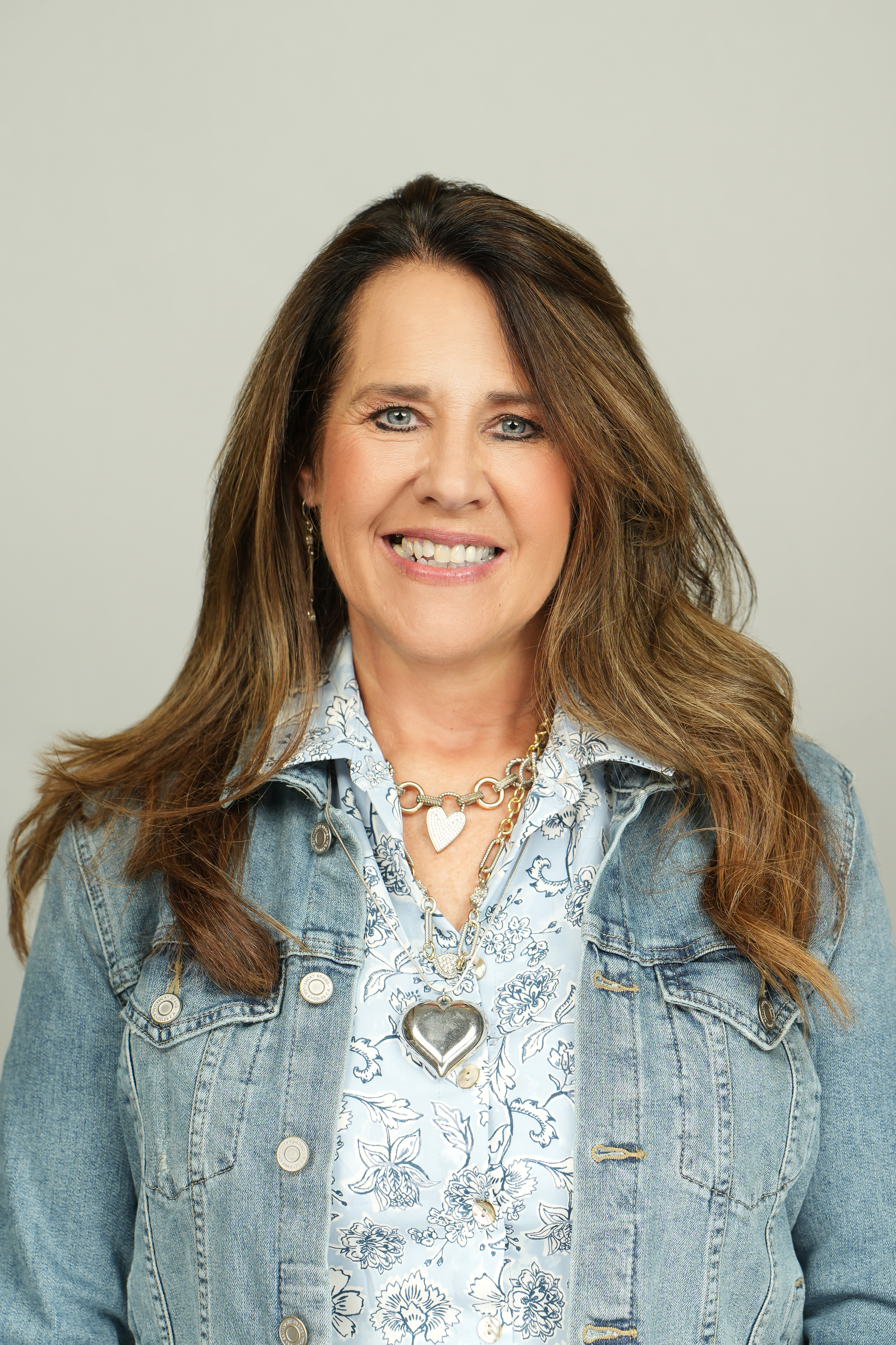 Professional headshot of a smiling person with long brown hair, wearing a denim jacket over a floral shirt.